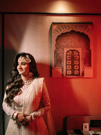 A beautiful portrait of the bride in her elegant white outfit, standing against a wall with warm, reddish lighting. Her serene expression and the artistic backdrop create a stunning image.