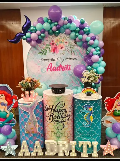 Aadriti's 7th birthday was an underwater adventure with this Little Mermaid theme. The setup includes character cutouts of Ariel, themed plinths with mermaid scales, and a balloon arch in shades of purple and teal.