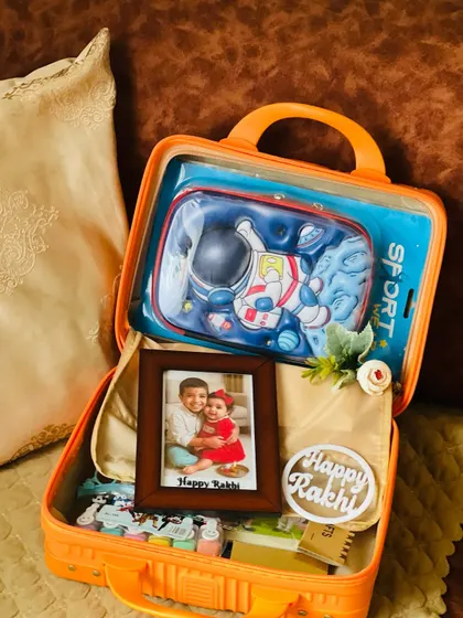 A special Rakhi hamper for a child, packed in a bright orange suitcase. It includes a personalized photo frame and an astronaut-themed pencil case to make the festival extra fun.