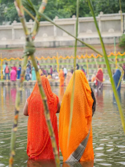 Two women in colorful sarees stand waist-deep in the water, offering prayers during Chhath Puja. The festival is marked by its simplicity and deep reverence for nature.