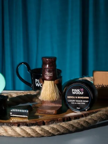 A collection of Pink Woolf men's grooming products styled on a rustic rope tray against a dark, moody background. This composition gives the brand a masculine and sophisticated aesthetic.
