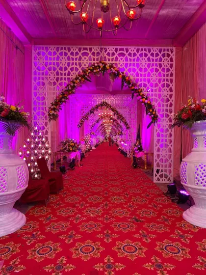 A grand and traditional entrance walkway with a red carpet, white lattice arches, floral arrangements, and rows of candle-style lights.