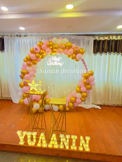 A stage setup with a pink and gold balloon ring against a white curtain backdrop. The decor is personalized with the name 'Yuaanin' in marquee letters.
