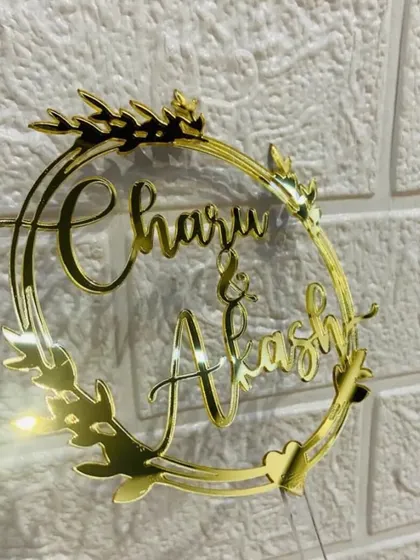The "Charu & Akash" topper with a leafy wreath design, shown against a lighted background to highlight its golden shine.