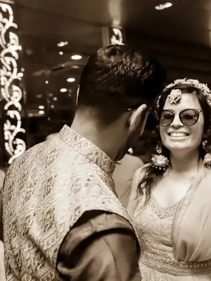 A candid, sepia-toned shot from a Haldi party. The bride, wearing sunglasses, shares a happy, knowing look with her partner, capturing the fun atmosphere.