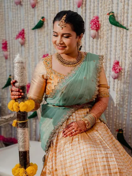 This bride looks radiant against a beautiful floral backdrop. The makeup is soft and traditional, perfectly matching her elegant half-saree and temple jewellery.