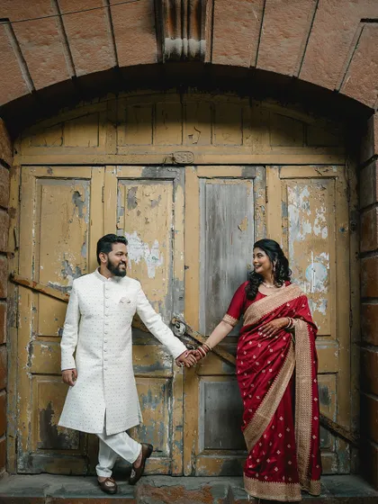 A couple holds hands, framed by a rustic yellow arched doorway. The contrast between her vibrant red saree and his white sherwani makes for a striking and romantic pre-wedding photograph.