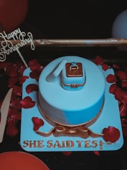 A special cake for a memorable proposal. This light blue cake is topped with a handcrafted, edible ring box and features "She Said Yes!" written in gold, set against a romantic backdrop of red rose petals.