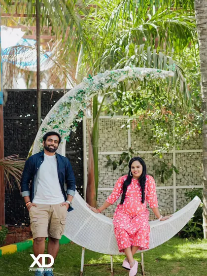 A whimsical couple's portrait featuring a large, flower-adorned crescent moon prop in a garden setting.