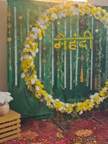 A Mehendi photo booth idea featuring a circular floral frame against a green draped background. The 'Mehendi' sign is made from marigold flowers.