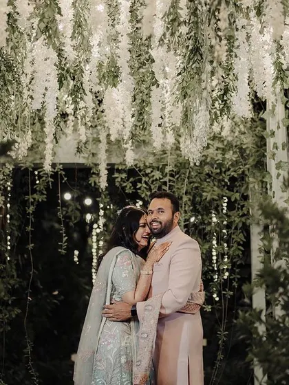 A cozy, romantic shot of a couple under a canopy of fairy lights and flowers. The warm lighting creates a magical atmosphere, perfect for a memorable Sangeet or reception portrait.