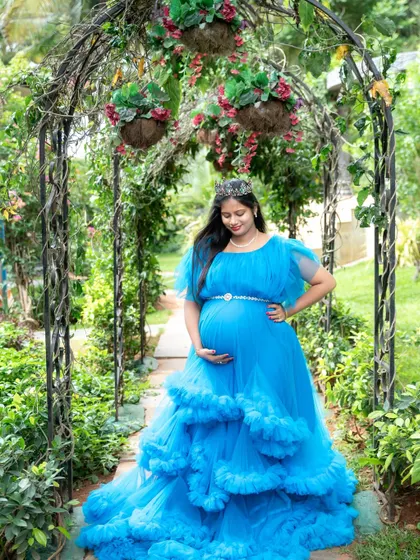 Feeling like a princess in a garden paradise. This mom-to-be wears a tiara and a stunning blue ruffled gown, standing under a floral archway.