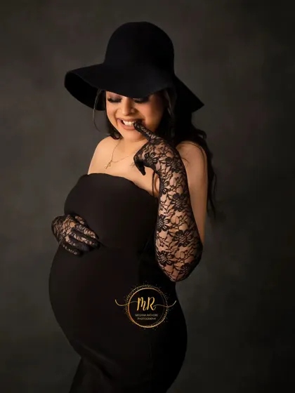 A cute and playful close-up portrait. The mother-to-be, dressed in a chic black hat and gloves, gives a joyful smile to the camera.