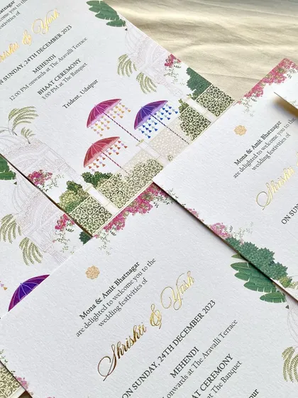 A pattern of Mehendi and Bhaat ceremony cards for the Udaipur wedding. The design combines colorful umbrellas, bougainvillea, and palm illustrations with elegant gold foil script.