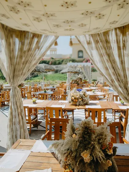 A view from inside a dining cabana, looking out onto the sunlit lawns. The long wooden tables are set with pampas grass centerpieces, creating a cohesive and natural aesthetic.