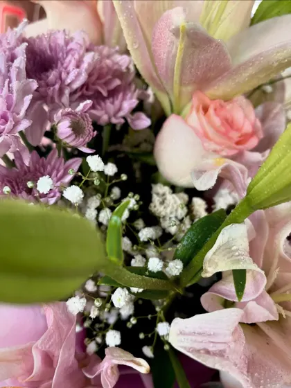A different angle of the fresh flower arrangement, showing the green leaves and baby's breath that add fullness and contrast to the pink and purple blooms.