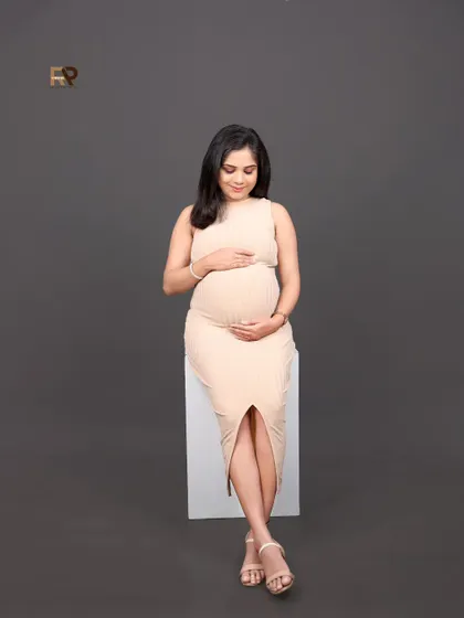 A serene solo portrait. The mom-to-be is seated, wearing a fitted beige bodycon dress that beautifully accentuates her baby bump. A perfect example of modern maternity elegance.