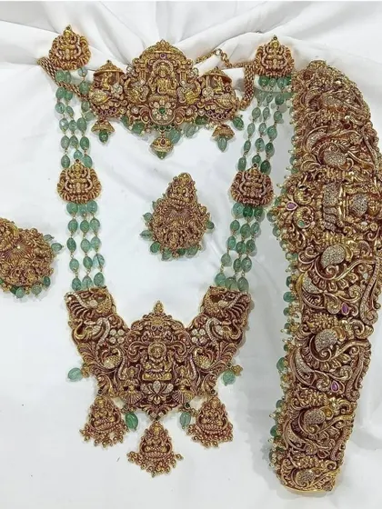 This is a magnificent antique bridal set featuring intricate carvings, goddess motifs, and green bead accents. It includes a choker, a long necklace, a statement vaddanam, and matching earrings.