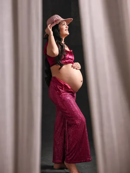 A chic and modern maternity look. The mom-to-be wears a stylish magenta sequin outfit with a matching hat, framed by curtains for a dramatic, fashion-forward portrait.