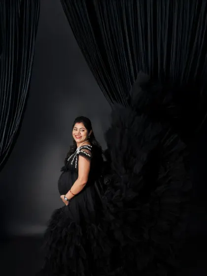 A dramatic and stylish solo portrait. The flowing black gown creates a powerful silhouette against the dark curtains, highlighting the mom-to-be's confident pose.