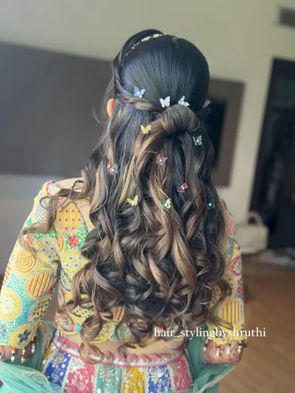 A playful and modern half-up hairstyle with voluminous curls, perfect for a Mehendi or Sangeet ceremony. The style is accented with whimsical multi-colored butterfly clips, adding a touch of personality and charm.