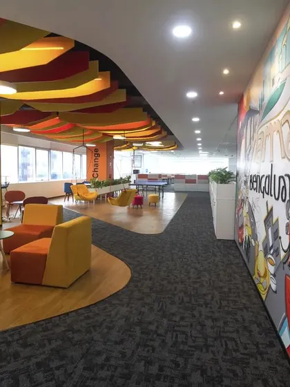 The long mural depicting Bengaluru's landmarks serves as a vibrant backdrop for this lounge area. The mix of armchairs and ottomans in warm colors creates a comfortable and visually rich environment for relaxation.