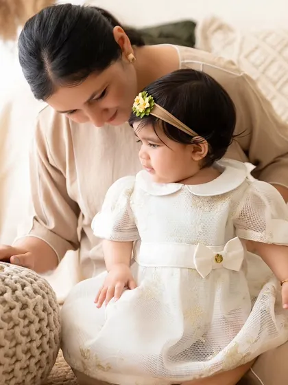 A mother interacts with her baby, who is sitting on the floor in a beautiful white dress.