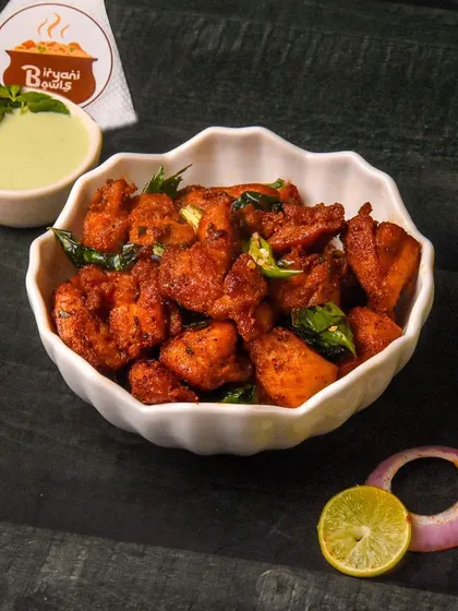 A bowl of spicy Chicken 65, a popular Indian appetizer, garnished with curry leaves and served with mint chutney. The dark background makes the dish look fiery and flavorful.