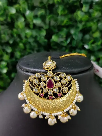 A stunning crescent-shaped maang tikka with intricate gold-tone engraving and a central red stone. The pearl drops add a touch of classic elegance.
