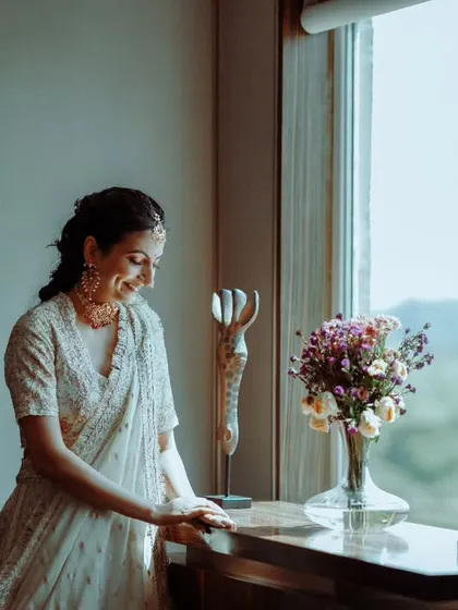 A candid moment of a bride or guest in a white outfit, smiling gently by a window.