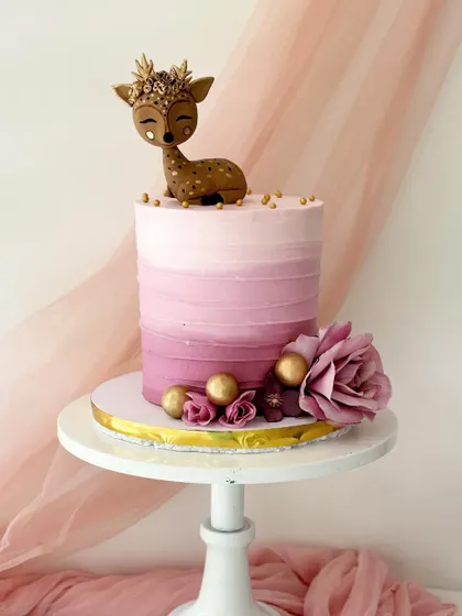 An elegant woodland deer cake with a pink ombre buttercream finish. The cake is topped with a hand-sculpted deer wearing a golden floral crown.