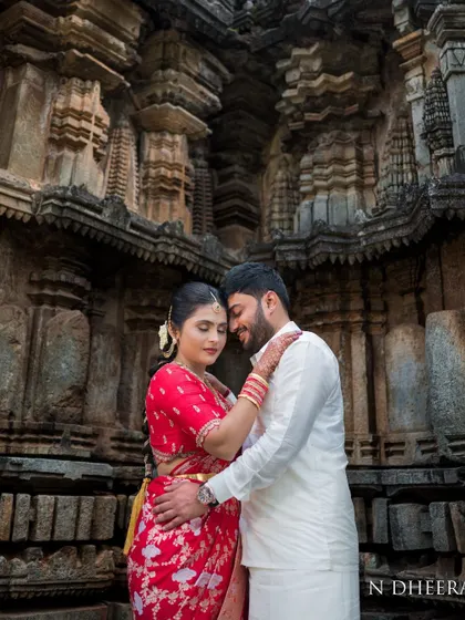 A romantic couple shot featuring the bride in her red saree and custom blouse. The traditional setting enhances the timeless quality of her outfit.
