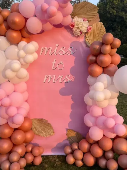 An outdoor bridal shower setup with a 'Miss to Mrs' backdrop. The balloon arch in shades of pink, white, and bronze looks beautiful against the natural green lawn.