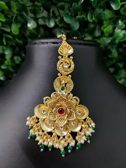 A beautiful temple-style maang tikka with a floral motif and antique gold finish. The green and red stones with pearl and bead drops make it a traditional favorite.