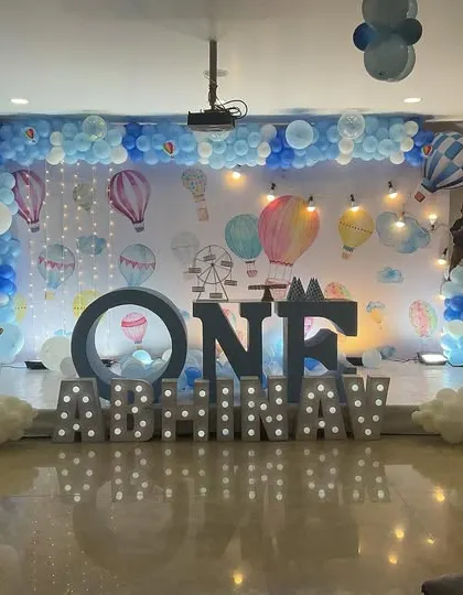 A grand hot air balloon themed first birthday stage decoration. The setup features large marquee letters spelling "ONE" and the child's name, a custom backdrop, and elaborate blue and white balloon arrangements creating a dreamy sky effect.