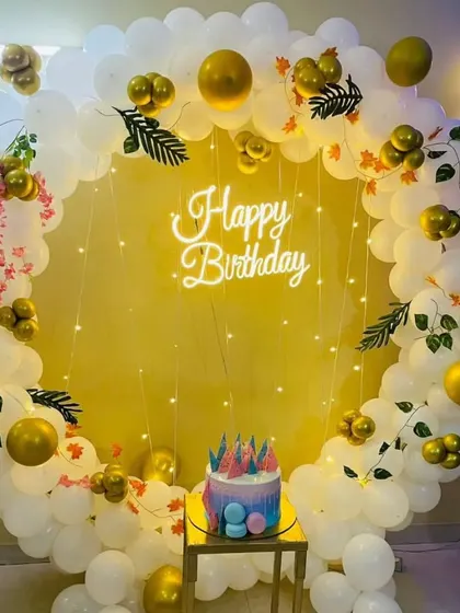 A birthday party with a circular balloon arch in white and gold. The decoration includes a "Happy Birthday" neon sign and a cake stand.