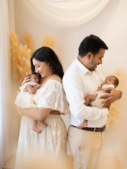 A slight variation of a classic pose, showing a different angle of connection. The parents stand back to back, holding their newborn twins and looking down at them with love.