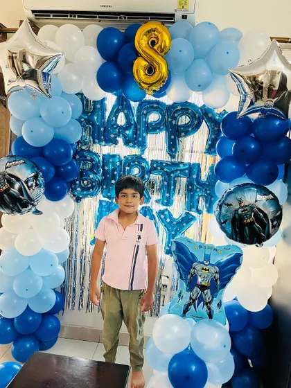 The birthday boy posing proudly in front of his Batman party setup. The decoration includes balloon columns and a "Happy Birthday" foil banner.