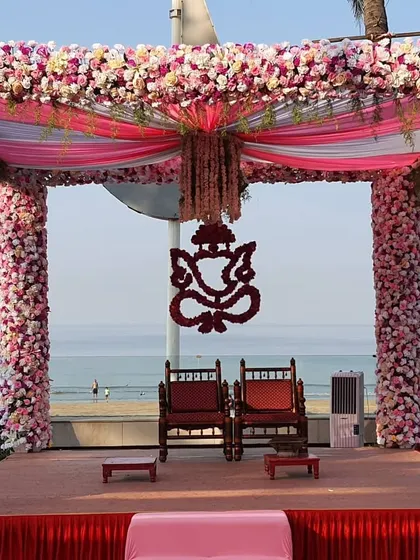 A close-up of a magnificent beachside mandap. The pillars are completely covered in pink flowers, and a floral Ganesh motif provides a traditional centerpiece, all set against the backdrop of the sea.