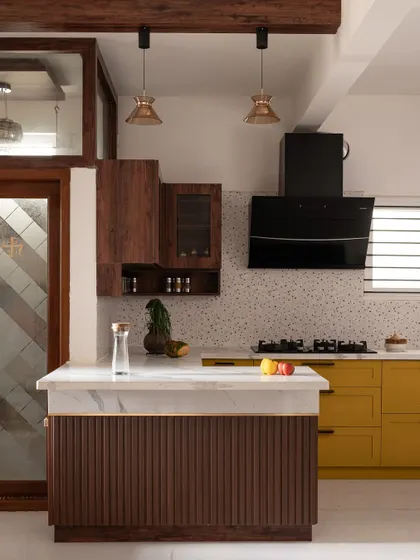 The fluted paneling on the breakfast bar and the glass pendant lights add texture and warmth to the 'Varnika' kitchen, complementing the bold yellow and rich wood tones.