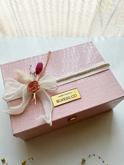 This pink leather-finish box is a sophisticated choice for a wedding invitation hamper. The exterior is adorned with a delicate ribbon and wax seal, hinting at the curated gifts waiting inside.
