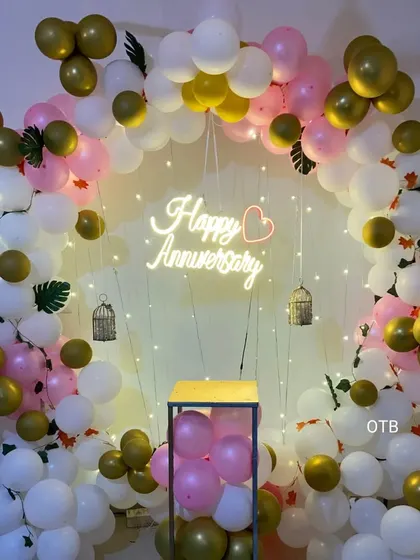 A beautiful and bright anniversary backdrop. This circular frame is decorated with pink, white, and gold balloons, fairy lights, and a glowing "Happy Anniversary" sign, perfect for taking memorable photos.