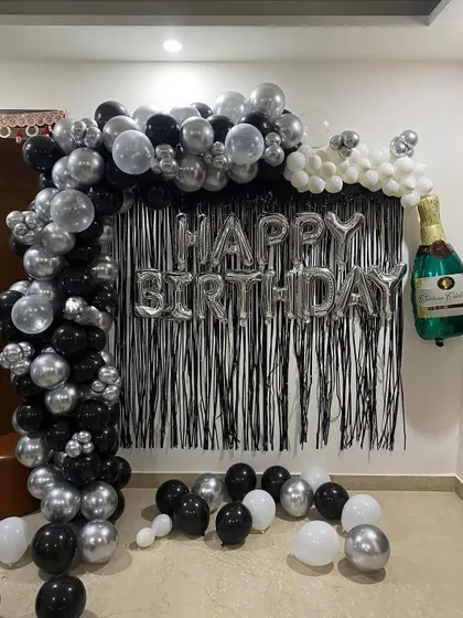A stylish black, silver, and white birthday decoration for a home party. The setup includes a balloon arch, a shimmer curtain, and a champagne bottle foil balloon.