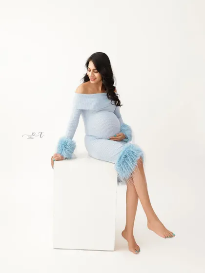 A chic and modern portrait. The mom-to-be is seated on a white block, wearing a stylish light blue dress with feathered cuffs, creating a fun and fashionable look.