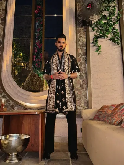 A full-length shot showing the complete black kurta and embroidered shawl ensemble. The outfit is styled for a festive night, combining traditional elements with a modern sensibility.