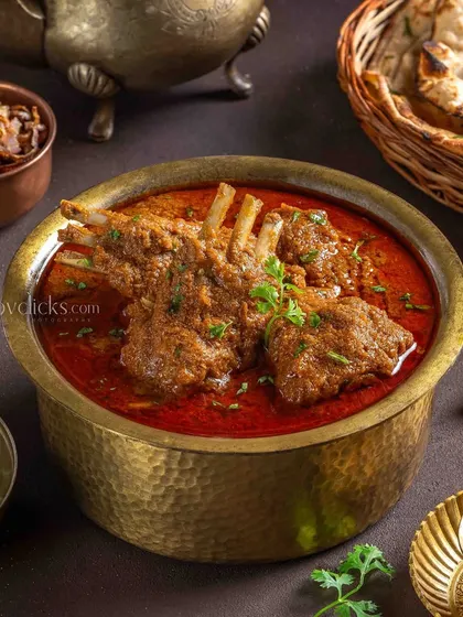 A rich and spicy Mutton Rogan Josh, with tender meat on the bone in a deep red gravy, styled with traditional brassware for an authentic feel.