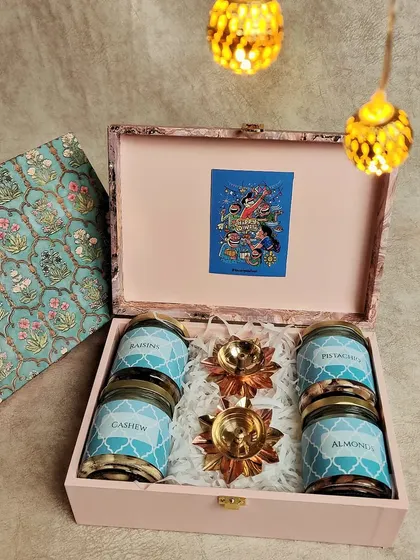 This beautiful wooden box hamper is perfect for Diwali. It contains four jars of assorted dry fruits and nuts, along with two decorative brass diyas, all nestled on a bed of white filler.