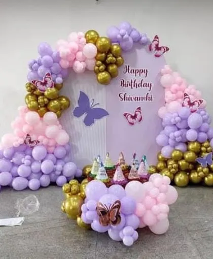 A pretty butterfly-themed decoration with a pink, purple, and gold balloon arch. The setup includes a custom backdrop and a small balloon arrangement for the cake table.