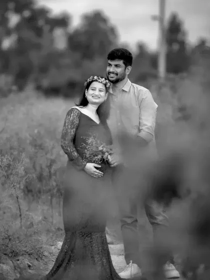 An artistic black and white outdoor maternity portrait. The couple stands together in a field, and the soft focus on the foreground flowers adds a dreamy, romantic feel.