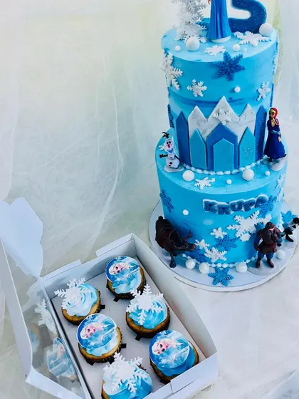 A stunning two-tier Frozen theme cake perfect for a winter princess. The cake is decorated with snowflakes, an ice castle, and beloved characters, paired with a box of matching themed cupcakes.